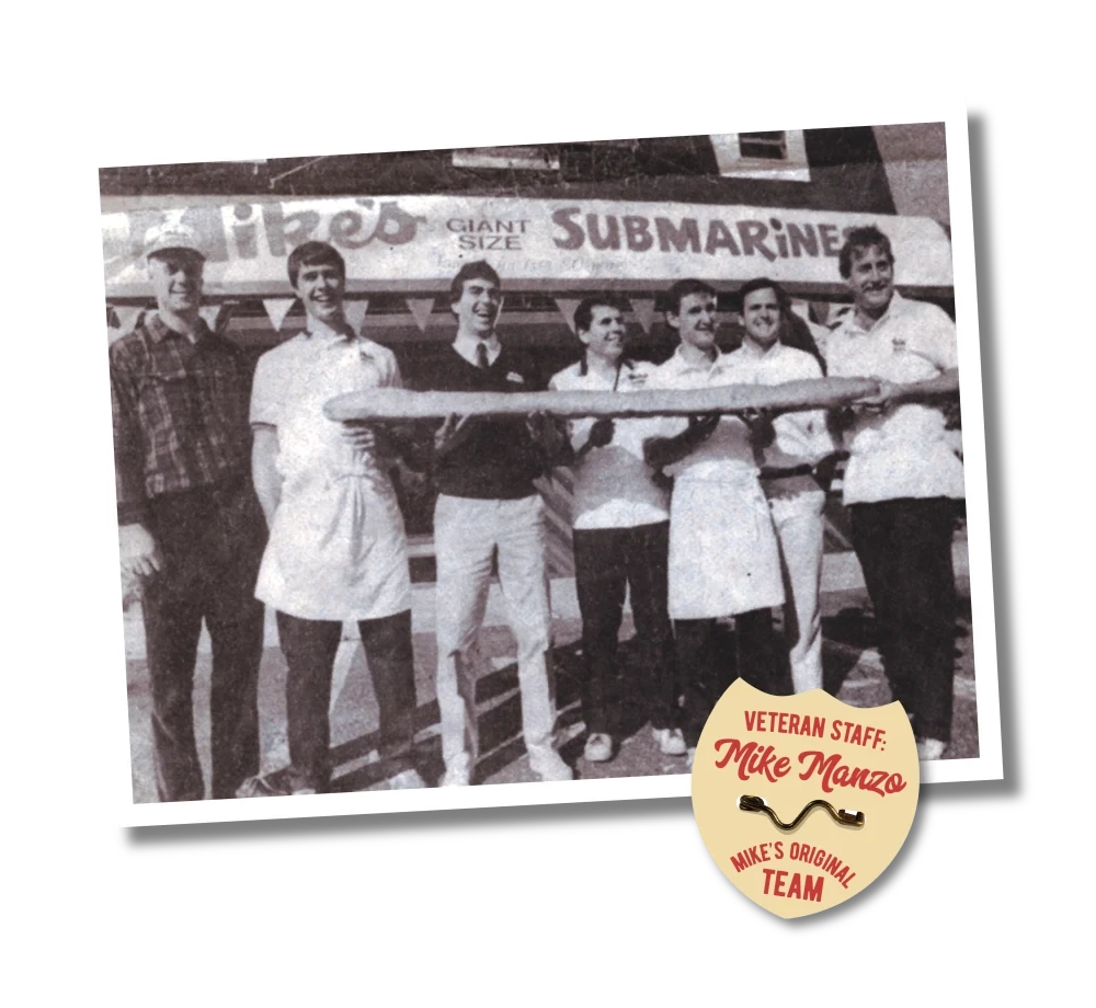 Sepia photograph of the original Jersey Mike’s staff outside the storefront, holding an oversized sub in front of the 'Giant Size Submarines' sign.